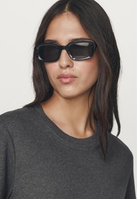 Occhiali da sole neri con una montatura spessa, forma leggermente arrotondata e lenti oscurate; il modello indossa un maglione grigio scuro a collo rotondo.