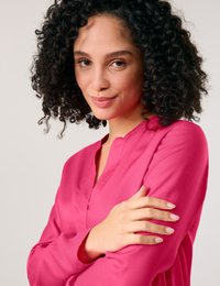 Fuchsia blouse with a V-neck design, long sleeves, and smooth texture. Model has curly hair and is crossing her arms.