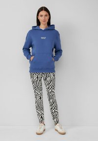 Blauwe hoodie met een voorzak, gecombineerd met zwart-wit zebraprint leggings en lichte sneakers. Gladde texturen door het geheel.