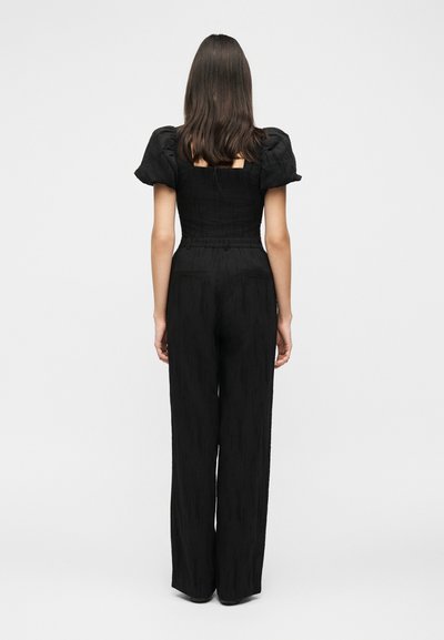 Γυναίκα με μακριά σκούρα μαλλιά στέκεται γυρισμένη πλάτη, φοράει ένα μαύρο ολόσωμο φόρεμα με φουσκωτά μανίκια και παντελόνια με φαρδιά ποδόγυρα σε απλό φόντο.