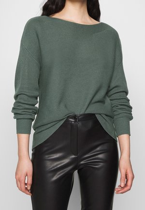 Žena v zeleném texturovaném svetr s dlouhým rukávem zasunutém do černých kožených kalhot s vysokým pasem na jednobarevném pozadí.