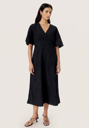Vestido midi negro con patrón de ojales en forma de diamante, escote en V, mangas acampanadas y cintura ajustada. Combinado con sandalias negras con tiras.