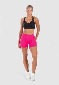 Schwarzer Sport-BH kombiniert mit leuchtend pinken Shorts. Die Shorts haben einen hohen Bund und eine glatte Textur. Das Model posiert vor einem einfarbigen Hintergrund.