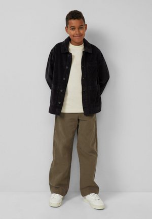 Zwarte corduroy jas met knopen, wit t-shirt, olijfgroene broek en witte sneakers. Lachend kind staat tegen een grijze achtergrond.
