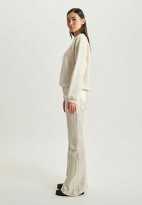 Pull léger beige avec un tricot texturé, associé à un pantalon évasé crème brillant. Le modèle se tient de côté, mettant en valeur les manches et l'ajustement du pantalon.