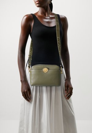 Green leather crossbody bag with gold hardware, featuring a front pocket and a logo detail, worn with a black top and white skirt.