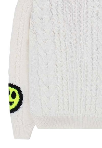 Maglione bianco lavorato a maglia con motivi a treccia, polsini a costine e un motivo di faccina sorridente nera e gialla fluo sulla manica sinistra.