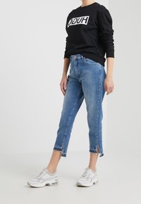 Persona que lleva puesta una camisa de sudadera negra con texto blanco en negrita en el pecho, jeans cortos azules con aberturas delanteras y zapatillas blancas.