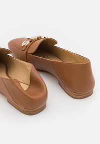 Mocassins en cuir tan avec des détails en métal doré, semelles plates en bois et un bout légèrement carré, présentés sur un fond blanc.