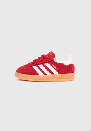 Red suede sneaker with white three-stripe design, textured heel, and gum rubber outsole. Features round laces and embossed logo on the side.