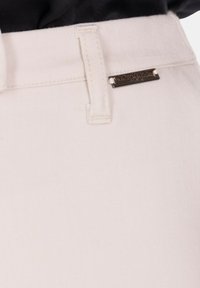 Primo piano della cintura di pantaloni beige chiaro con passante per cintura e una piccola targhetta metallica del marchio con la scritta "Mimi-Muà Paris".