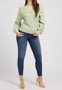 Femme portant un pull léger vert d'eau à épaules dénudées, un jean skinny bleu et des sandales à talons jaunes, se tenant devant un fond uni.