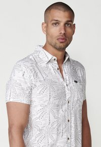 Camisa de manga corta en tejido blanco claro con un patrón de hojas de palmera en tonos grises, collar abotonado y un pequeño bolsillo en el pecho con branding.