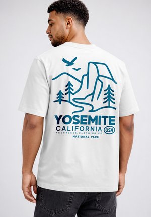Man wearing white shirt with blue Yosemite National Park graphic showing mountains, trees, birds, and text "California USA" on back.