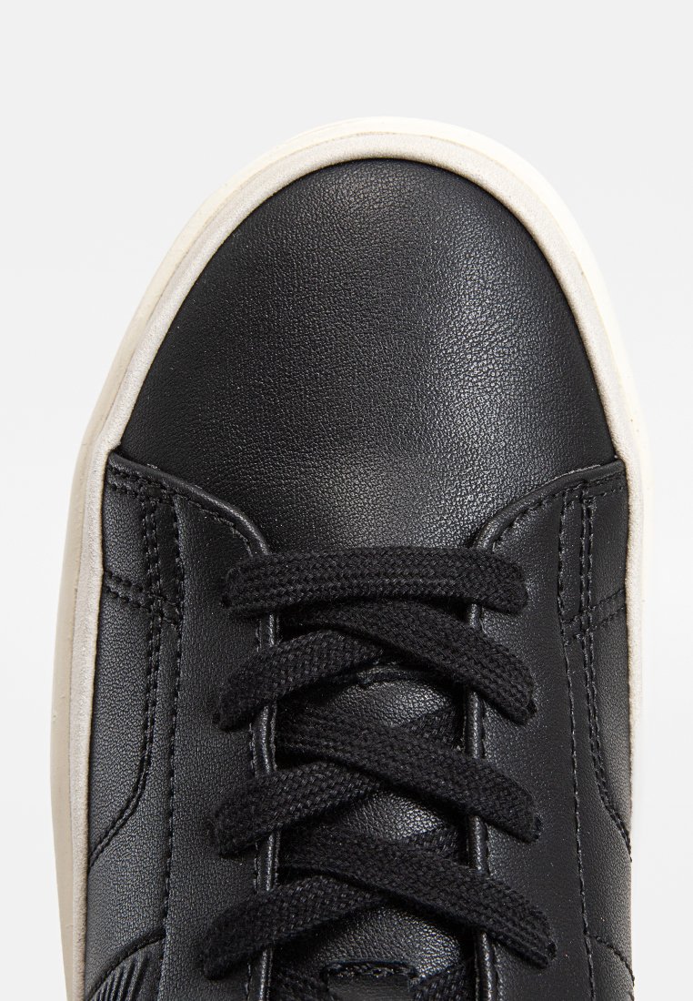 Zapatilla de cuero negra con puntera redondeada, cordones negros y suela de goma blanca. Costuras visibles y detalles de superficie texturizada.