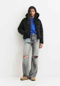 Zwarte pufferjack met capuchon, blauwe trui eronder, gestreept shirt dat eruit piept, versleten lichtgrijze wijde jeans en beige schoenen.
