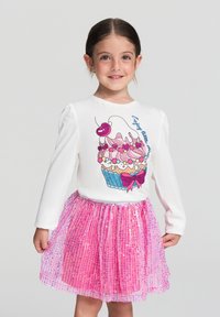 Biała bluzka z długim rękawem z grafika babeczki, błyszcząca różowa spódniczka z tiulu i pasem. Spódnica ma pionowe cekiny dla dodatkowej tekstury.