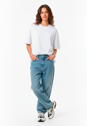 Tricou alb din bumbac, cu mâneci scurte, asortat cu blugi de denim bleu deschis. Purtați cu adidași negri și albi, cu mâinile în buzunare.