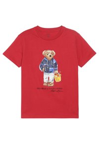 Camiseta roja de manga corta con un gráfico de un oso con chaqueta azul y pantalones blancos, sosteniendo un cubo amarillo, con una marca impresa debajo.