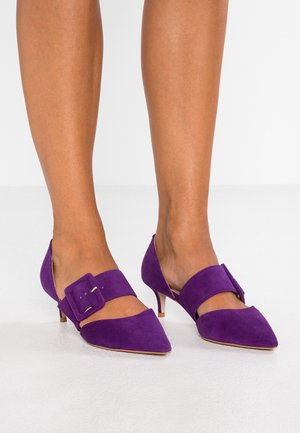 Ben iført lilla ruskind kitten heels med spidse snuder og brede spænder, stående på et hvidt gulv mod en ensfarvet baggrund.