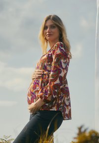 Kleurrijke blouse met paisleypatroon en lange pofmouwen, gedragen over donkere jeans. De stof is licht en vloeiend, wat de silhoutte benadrukt.