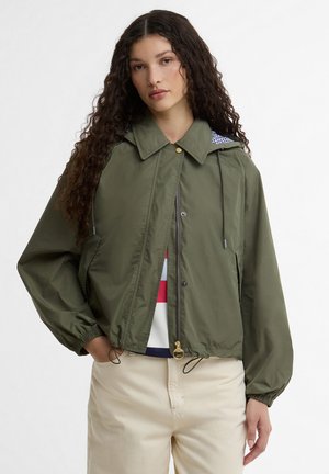 Fiatal nő hosszú, göndör hajjal, olívazöld kapucnis dzsekit, csíkos inget és bézs nadrágot viselve, egyik kezét a zsebében tartva áll.