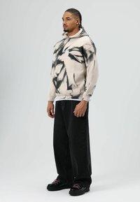 Beige hoodie met zwart tie-dye patroon, voorzak, gecombineerd met losse zwarte jeans en chunky zwarte sneakers met roze veters.