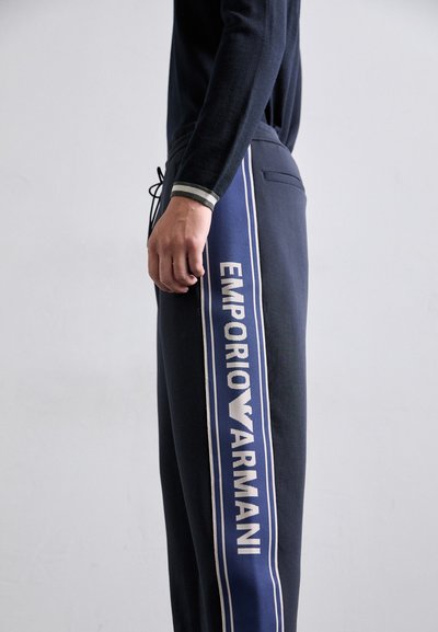 Persoană îmbrăcată în pantaloni de trening bleumarin cu o dunga laterală și textul "EMPORIO ARMANI" în alb pe dungă, pe un fundal simplu deschis.