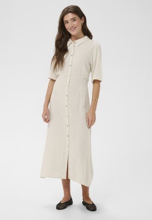 Junge Frau lächelt, trägt ein beiges Midikleid mit kurzen Ärmeln und Knopfleiste sowie schwarze gewebte flache Schuhe vor einfachem Hintergrund.