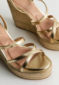 Sandalias de cuña metálicas doradas con correas entrecruzadas, que cuentan con una plataforma de yute trenzado y un forro interior suave de color rosa claro.