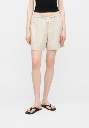 Person iført beige shorts med snøre, sort ærmeløs top og sorte flip-flops, stående mod en simpel lys baggrund.