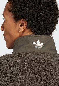 Braune Fleecejacke mit strukturiertem Obermaterial und hohem Kragen. Besitzt ein weißes Logo am Kragen und einen glatten Stoffbesatz am Hals.