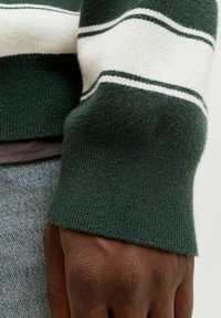 Zaļa adīta džemperu piedurkne ar baltām un tumši zaļām svītrām. Mīksta tekstūra, ribota apdare, daļēji redzama roka un izbalējušas zilas džinsu drēbes fons.