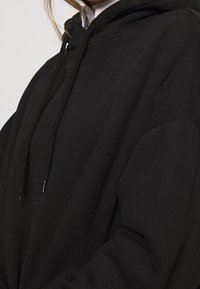 Felpa nera con cappuccio regolabile, realizzata in morbido tessuto. Vestibilità ampia con spalle scese e polsini a coste. Design minimalista, senza loghi visibili.
