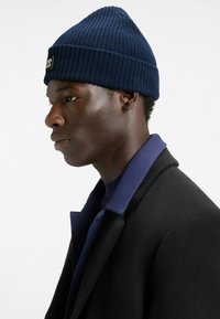 Navy ribbed knit beanie with a folded cuff. Worn with a black coat and a purple shirt underneath. Smooth texture, close-fitting design.