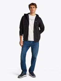 Haut à capuche noir zippé avec cordons de serrage, T-shirt blanc avec logo, jean bleu et baskets sombres. Tenue décontractée avec une coupe ample.