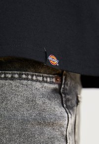 Crna pamučna majica kratkih rukava s zaobljenim rubom, s malom crvenom i žutom oznakom "Dickies" blizu donjeg šava, uparena s sivim trapericama.