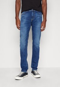 Blå denimjeans med slim fit, med kontrasterande sömmar, femfickorsdesign och dragkedja. I kombination med svarta sneakers.