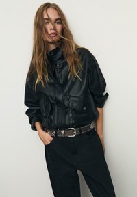 Černá kožená krátká bunda s vysokým límcem a dvěma předními kapsami, v kombinaci s tmavými kalhotami a hnědým cvočkovaným pásem. Hladká textura.