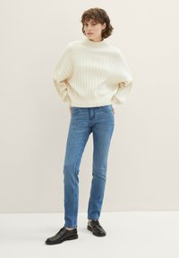 Kreemjas soonikuga sviiter, millel on kõrge kaelus ja lõdvas stiilis, koos helesiniste kitsaste teksade ja mustade nööbiga kingadega.