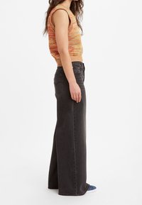 Woman wearing sleeveless multicolor knit top, high-waisted wide-leg black jeans, and blue shoes, standing in profile on white background.