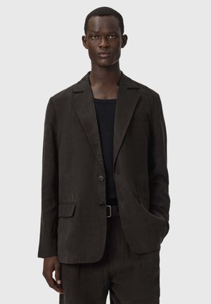 Junger Mann, der einen dunkelbraunen Blazer über einem schwarzen Hemd trägt, mit einer Hand in der Hosentasche, vor einem schlichten hellen Hintergrund stehend.