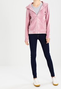 Kvinna klädd i en ljusrosa zip-up hoodie, grå t-shirt, mörka tajta jeans och vita sneakers med bruna sulor, stående mot en enfärgad bakgrund.