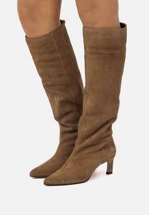 Botas marrones de ante hasta la rodilla con punteras puntiagudas y tacones bajos, usadas en las piernas sobre un fondo blanco liso.