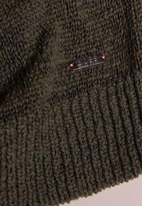 Stickad mörkgrön mössa med ett strukturerat mönster, ribbade kanter och en liten metall-logotag. Mjuk material med en elastisk passform.