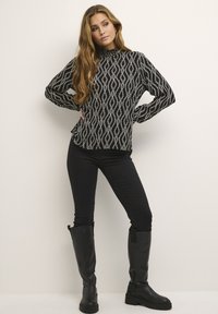 Blouse noire à motifs avec des formes de diamant blanches, col montant, manches longues, associée à un jean skinny noir et des bottes noires montantes.