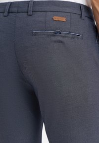 Marineblaue, strukturierte Hose mit einer Gesäßtasche, die über einen kontrastierenden Knopf und ein Lederetikett mit der Aufschrift "bugatti" verfügt. Dezentes diagonales Muster.