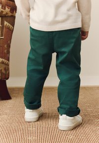 Pantalons en coton verts avec les poignets retroussés, associés à des baskets blanches, sur un tapis beige texturé et une chaise en tissu à motifs.