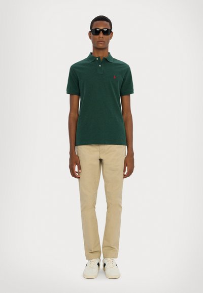 Mörkgrön pikétröja med korta ärmar, khaki chinos och vita sneakers. Modell med mörka solglasögon står mot en enkel bakgrund.