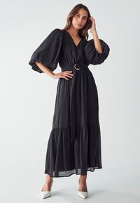 Black maxi dress with pleated texture, V-neckline, puff sleeves, and a tied waist with a round belt. Worn with white heeled shoes.